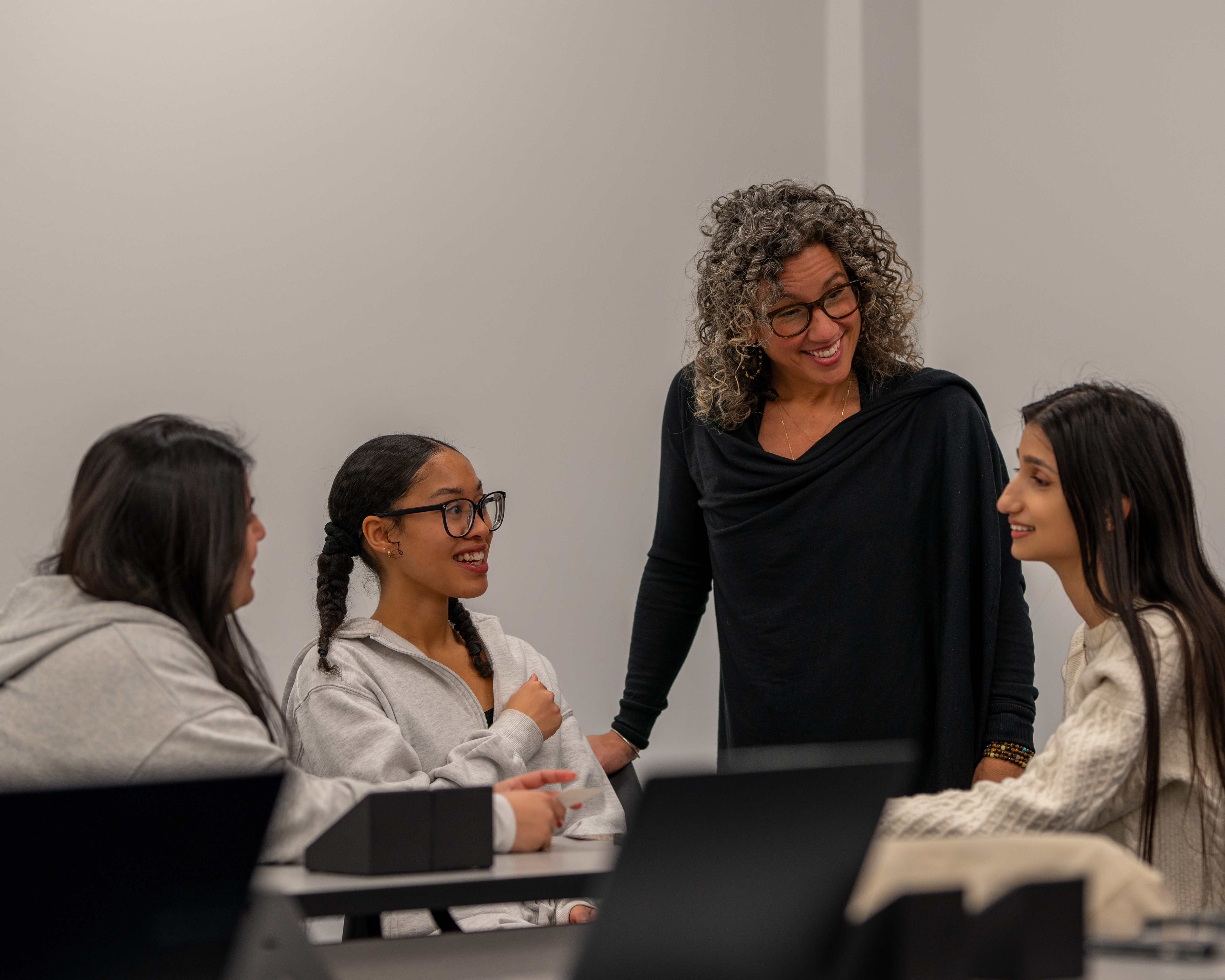 A female professor engages warmly with three students during a classroom discussion. Everyone is seated around a table with laptops, appearing focused and collaborative. 